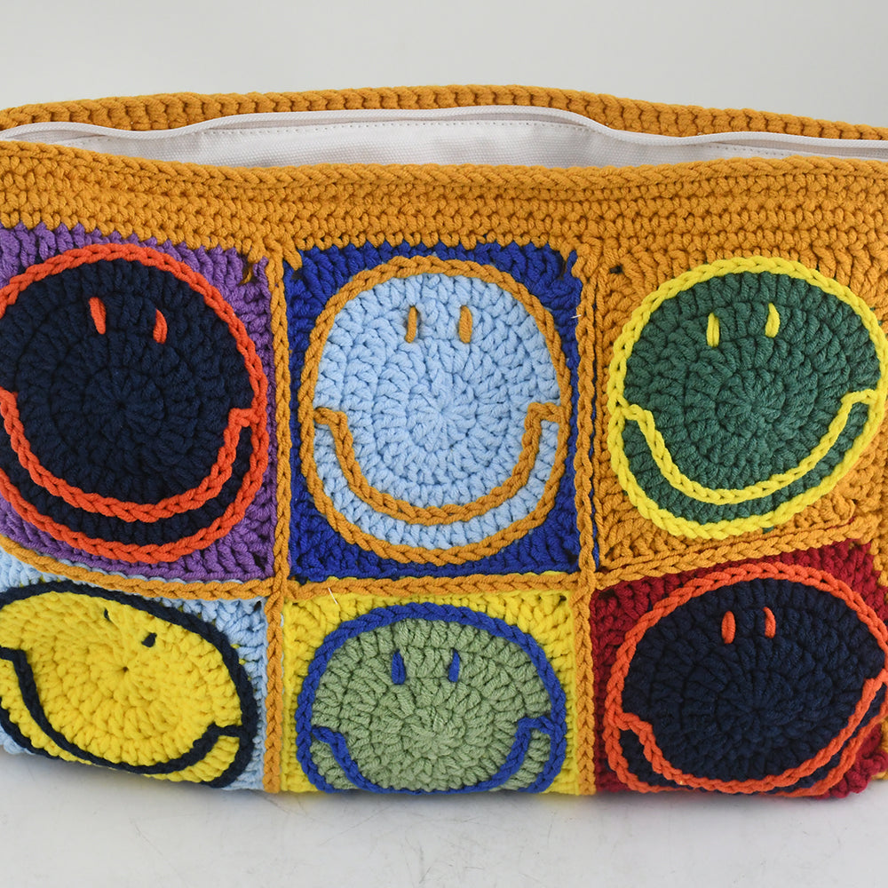 Handmade crochet clutch bag featuring colorful smiling face motifs in pink, yellow, blue, green, red, and orange squares. Lightweight, eco-friendly, and compact design with a secure zipper closure.