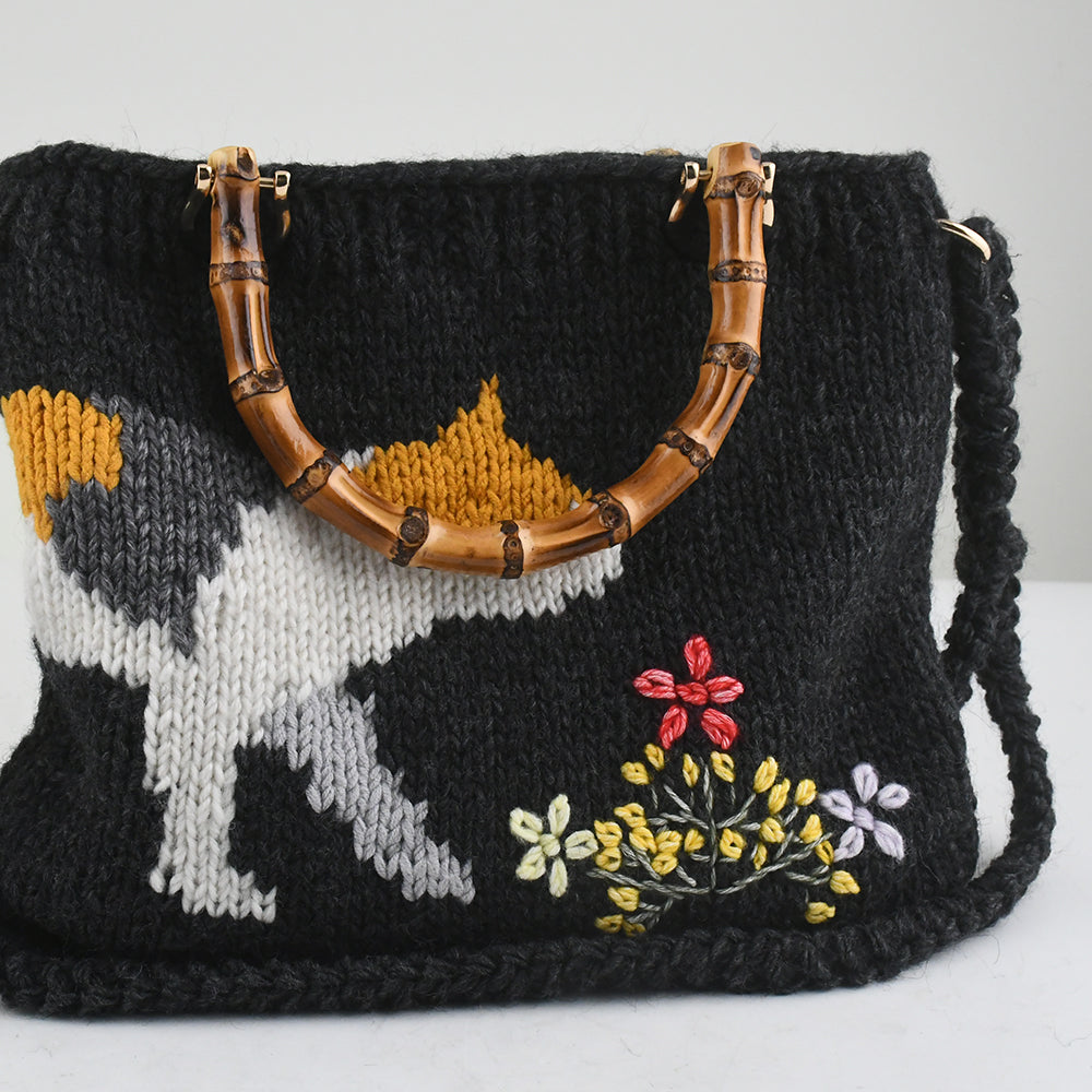 Handmade knitted black tote bag featuring a bamboo handle, embroidered cat design, and colorful floral accents. Lightweight and eco-friendly, perfect for casual or creative fashion styles.